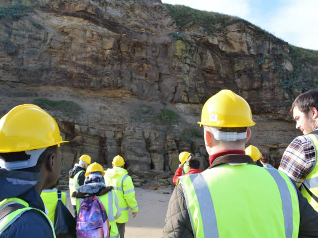 Students wearing high-vis jackets and hard hats examining a rock face in Whitby.