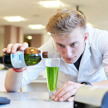 A pharmacy student formulating medicine by pouring a solution from a bottle into a glass of green liquid