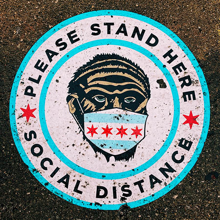 Sign on the floor with a graphic of a person wearing a mask and the text 'Please stand here, social distance.'