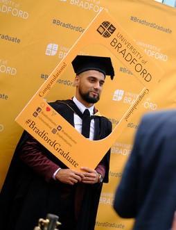Graduating student in cap and gown posing for a picture with #BradfordGraduate prop