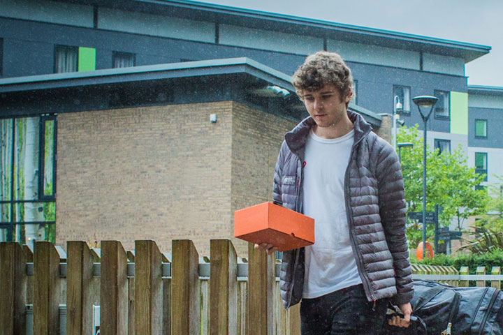 A student walking in the rain holding a small red box and pulling luggage.