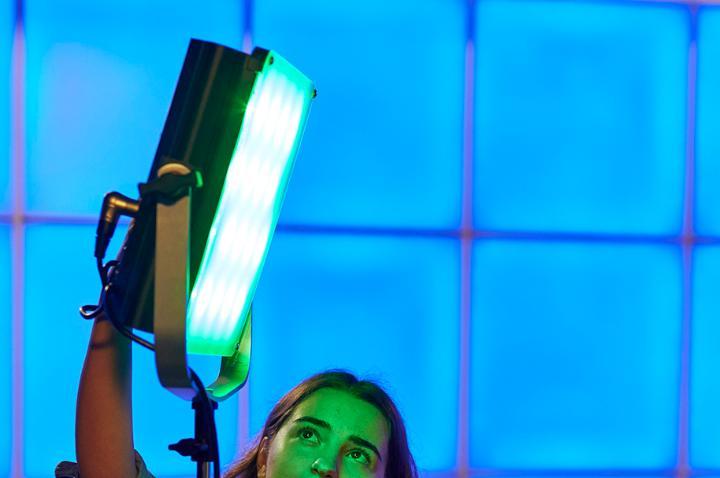 A large bright green spotlight light in a TV studio. The walls are blue. At the bottom of the frame, a student, whose face is illuminated by the green light, is adjusting the angle of the spotlight.