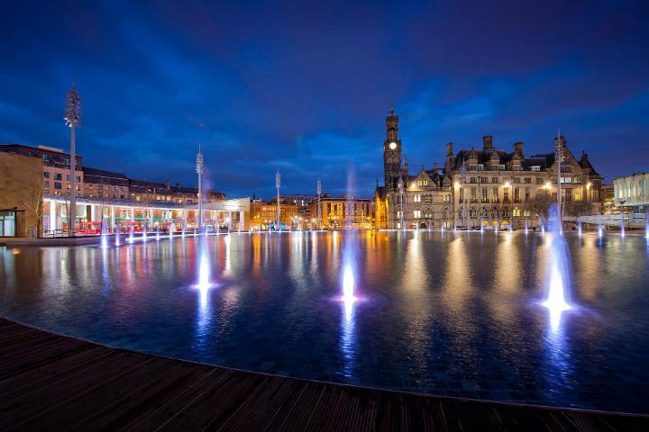 An evening photo of Centenery Square in the centre of Bradford, featuring a water fountain and City Hall in the background.