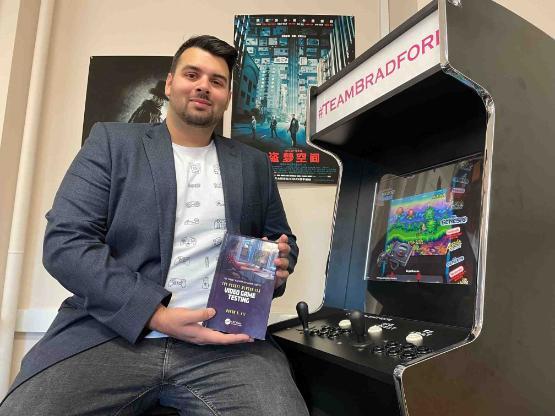 Former student sat down holding a book next to games arcade machine
