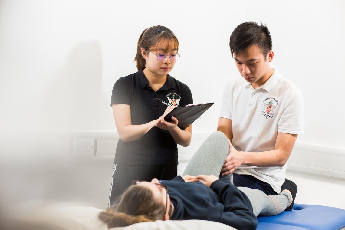 A physiotherapy student bends a patient's leg while being assessed by another physiotherapist