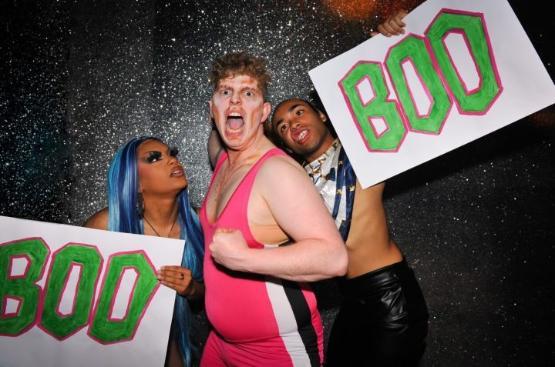 Three wrestlers in costume hold up large signs with Boo written on them