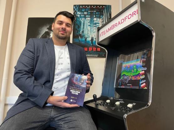 Former student sat down holding book next to video game arcade machine