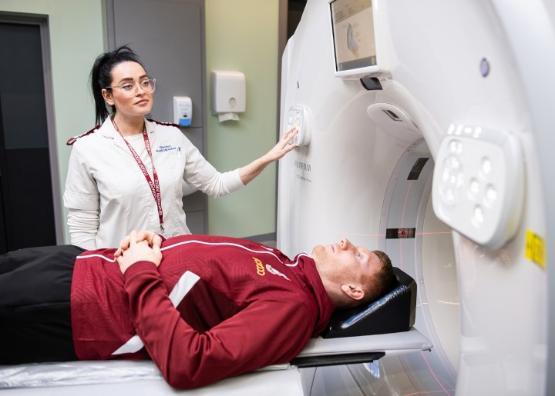A football players is laying down and about to be put through a CT scanner machine supervised by a student