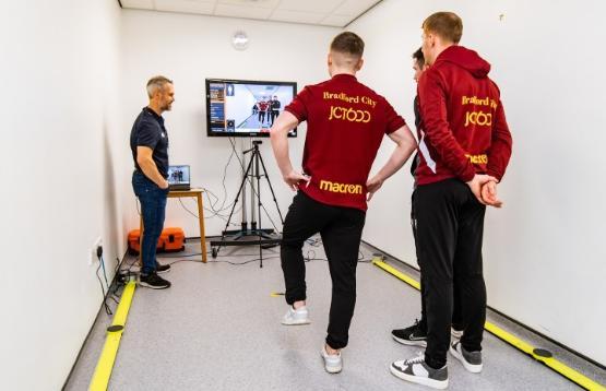 Three football players standing up in a physiotherapy room using the equipment looking at a large TV in front of them