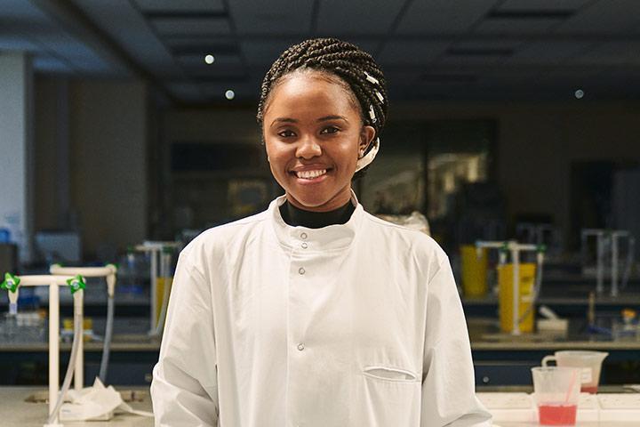 Student in a lab coat. They are in a lab setting and smiling at the camera.