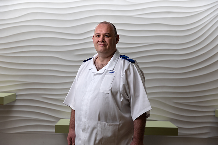 Student nurse, Chris, in uniform stood in front of a white textured wall