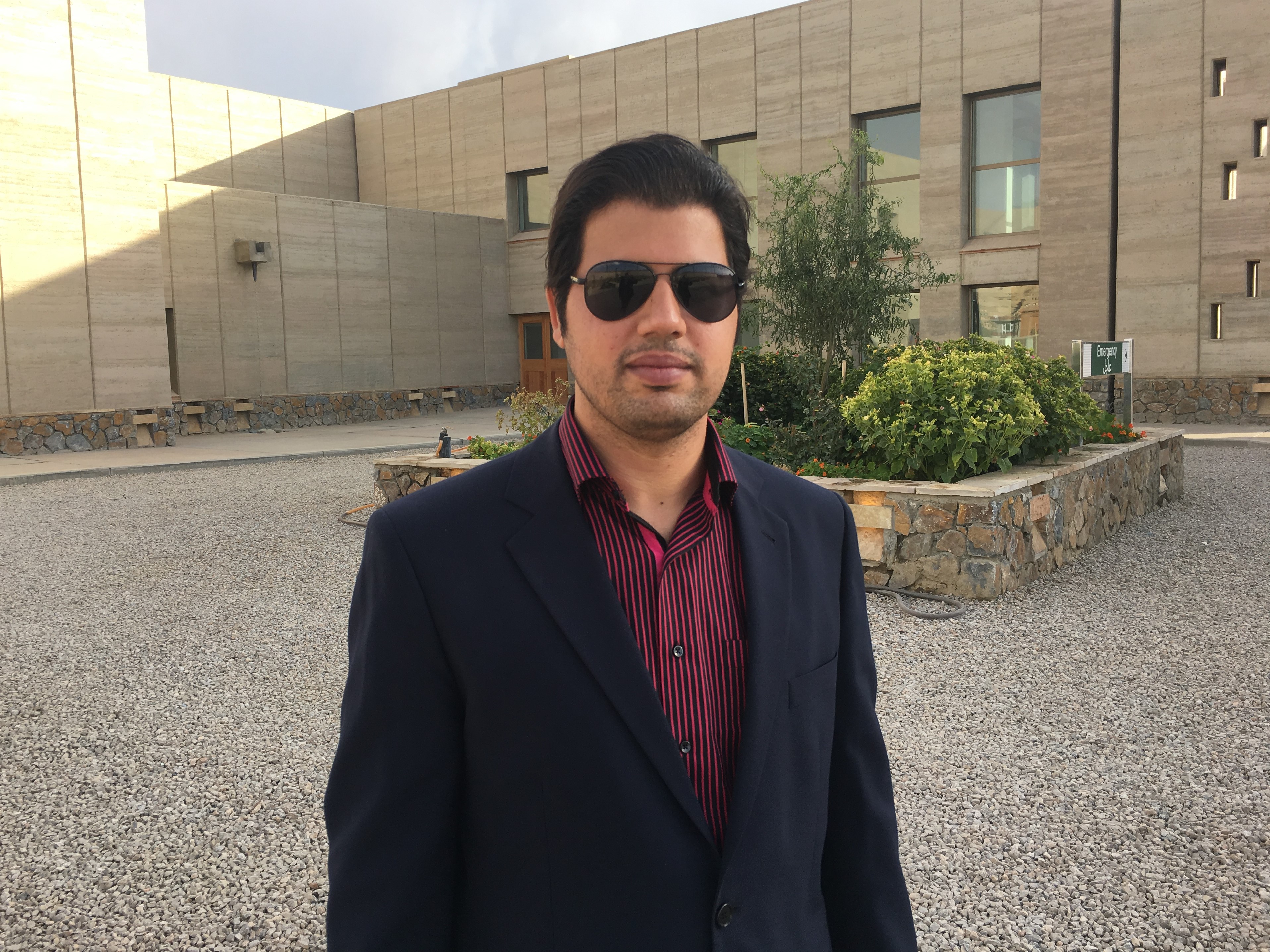 Fahim wearing a suit jacket, shirt and shades, stood in front of a flower bed.