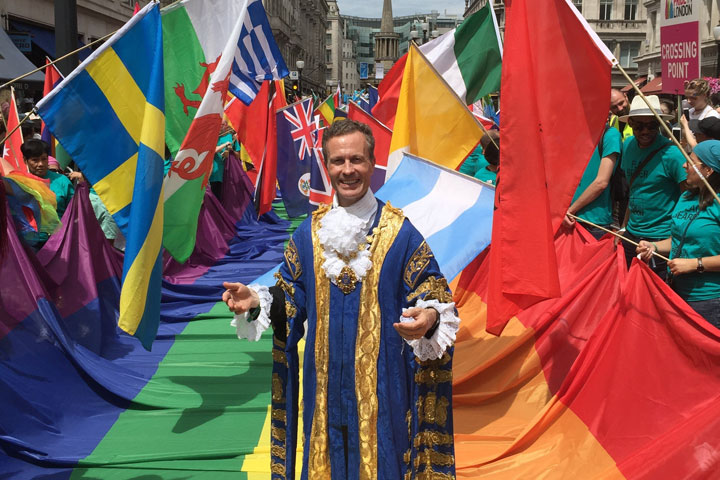 A smiling person wearing a blue and gold gown, surrounded by colourful flags and several people.