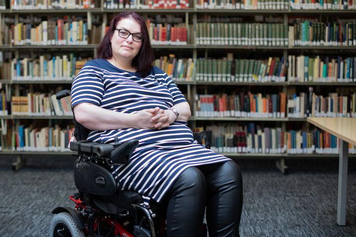 A person in a library, sitting in a wheelchair. Shelves full of books are in the background.