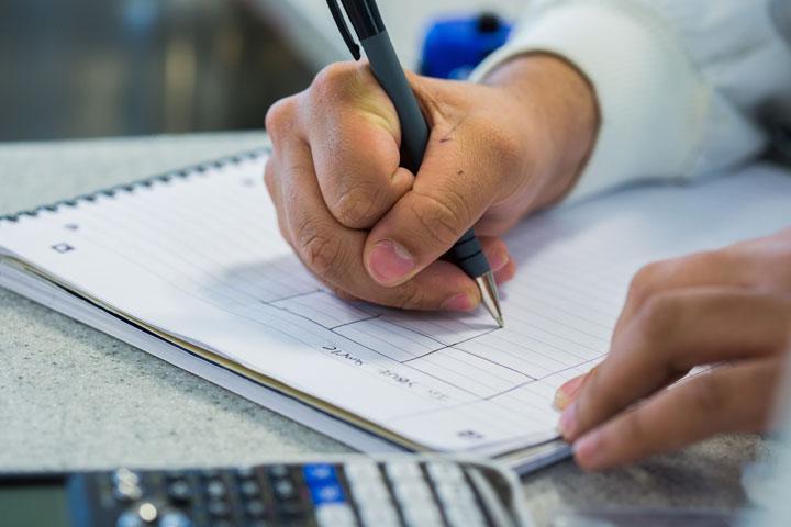 A close-up of a hand holding a pen and writing on a notepad. There is a calculator on the desk beside the notepad.