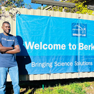 A smiling person stood in front of a blue sign that reads, Welcome to Berkeley Lab.