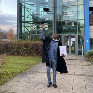 Graduate Billy wearing his gown and tossing his cap in the air outside the Institute of Life Sciences Research building