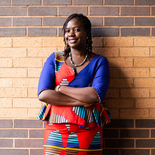 A person wearing brightly coloured clothing leaning against a wall with arms folded, smiling.