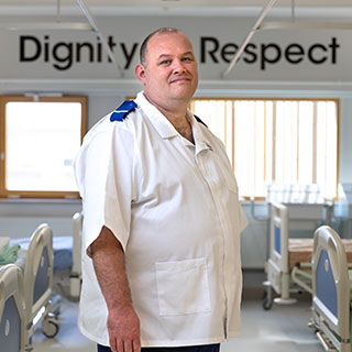 Student stood in a nursing uniform in a clinical setting. They are stood in front of a sign that reads 'Dignity, Respect'. 