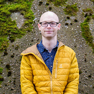 A student wearing a yellow coat standing in front of a wall of moss smiles at the camera
