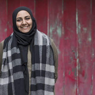 Bradford student Maria, stood in front of a red wooden garage door, wearing a black headscarf and a checkered scarf.